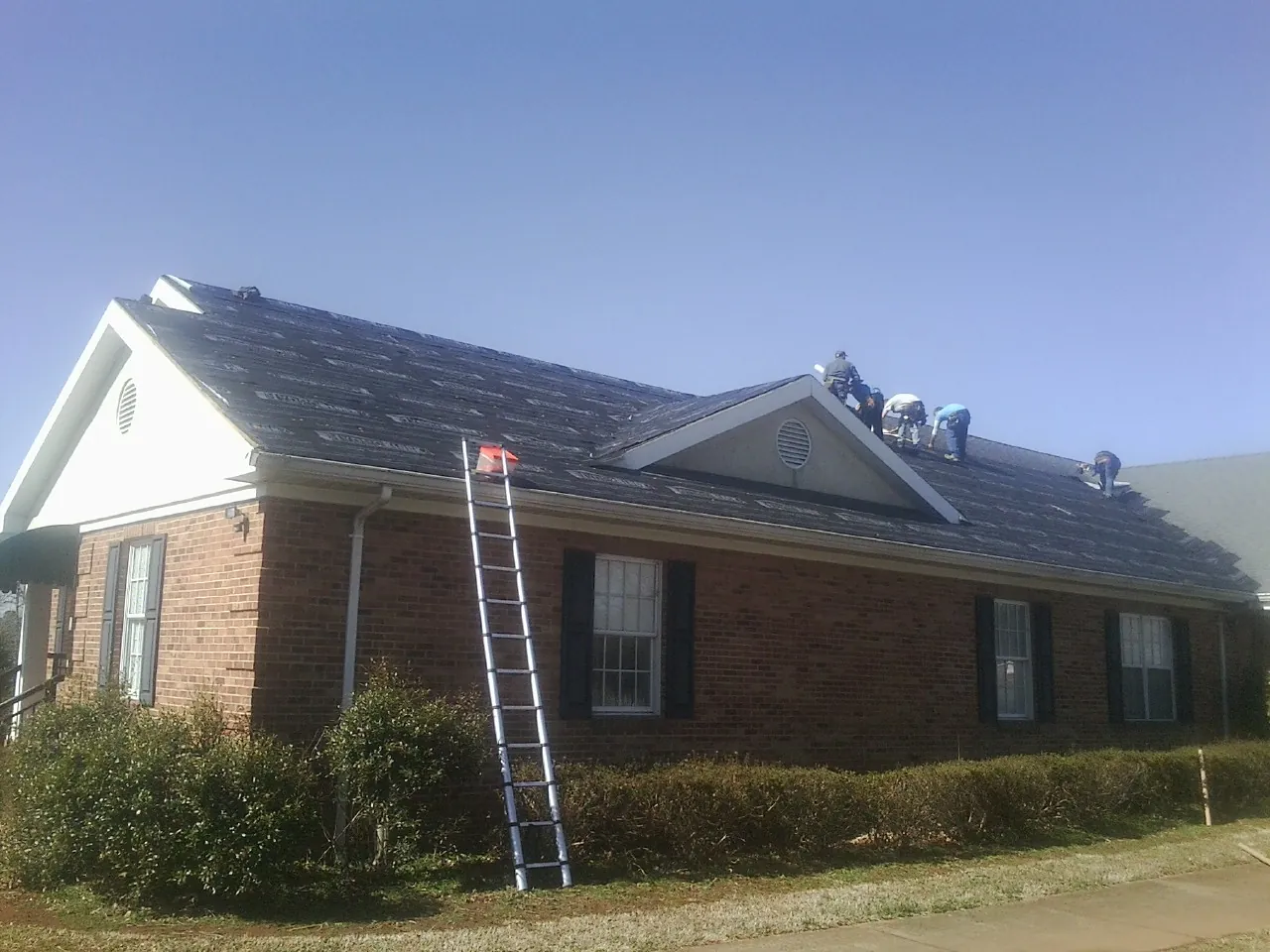 Crew tearing shingles off a brick ranch roof with a ladder set — Emergency Roof Repair work by our crew in Hestand