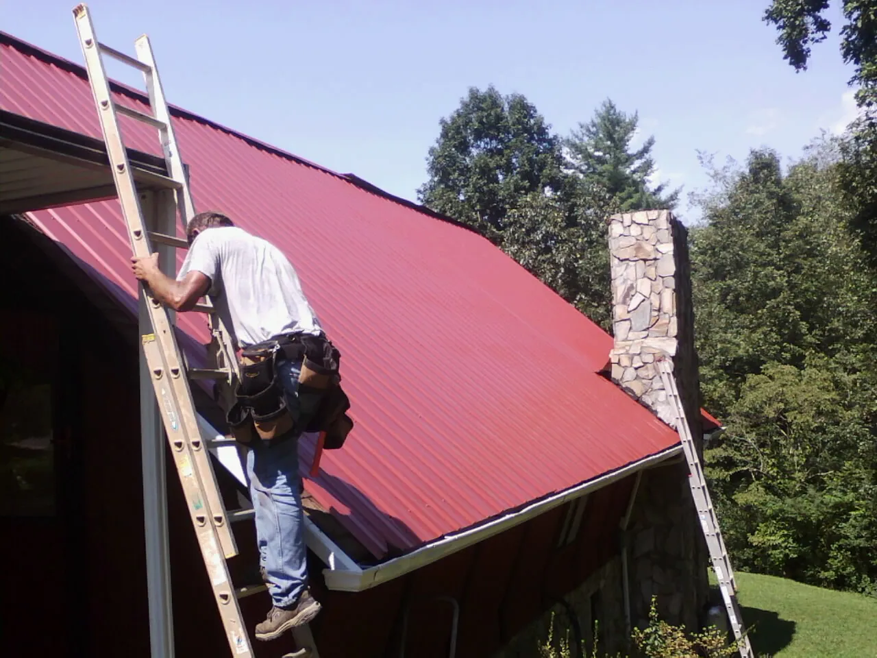 Roofer on a ladder beside a red metal roof and stone chimney — Soffit & Fascia Repair by Roofing Brothers in Hestand