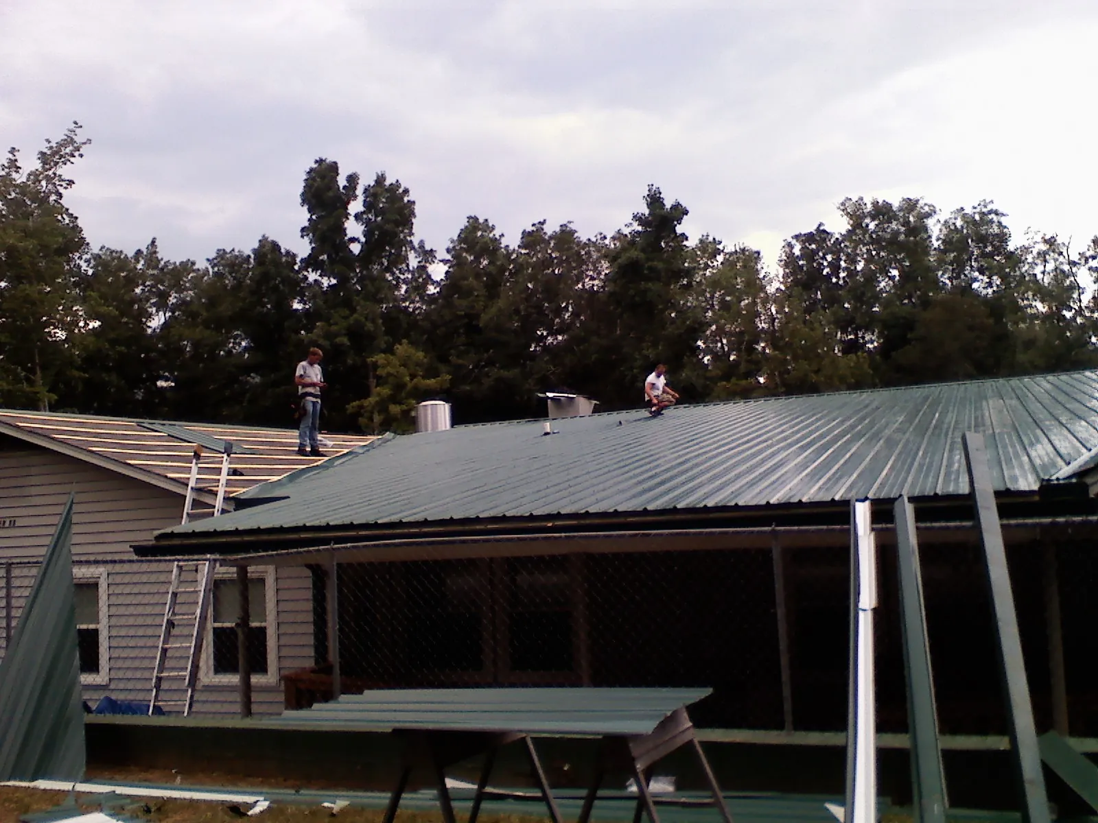Two roofers installing green metal panels above a covered porch in Hestand